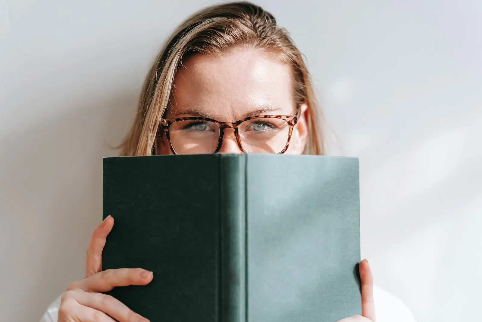 A woman's face partially hidden behind a book with a green cover.