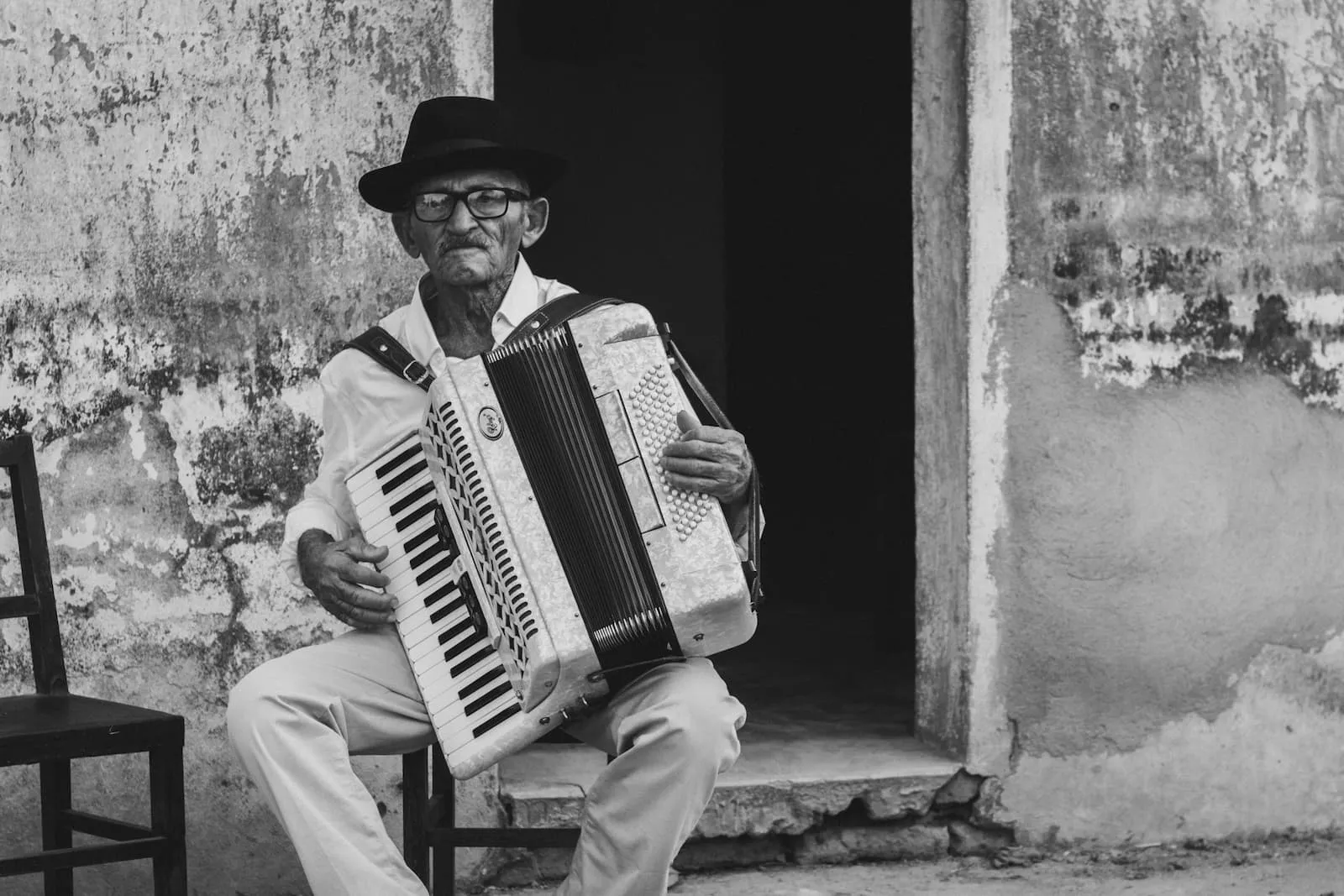 An older man sitting on a chair in front of a doorway, playing accordion.