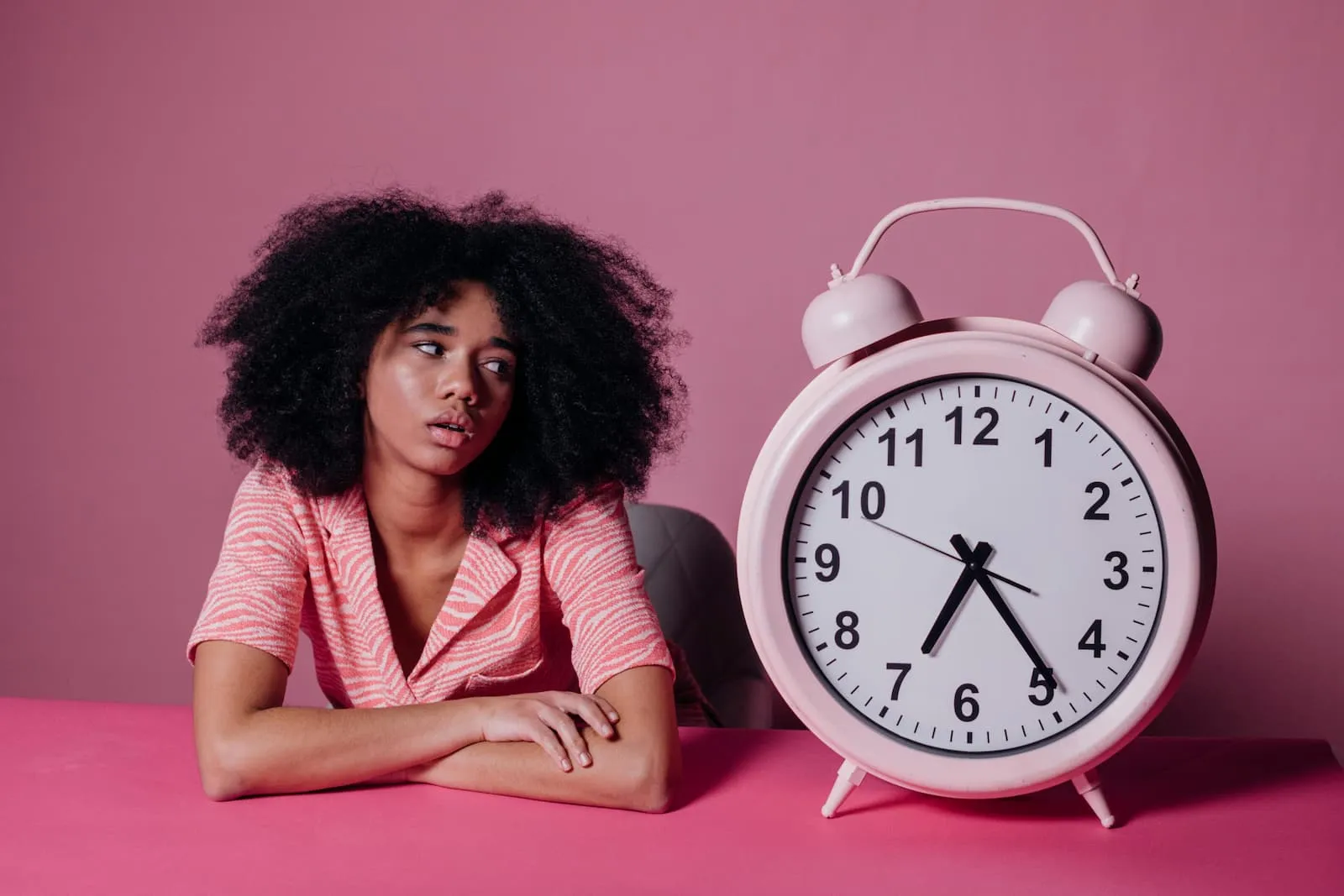 A woman rests her arms on a table, looking at the big alarm clock next to her.