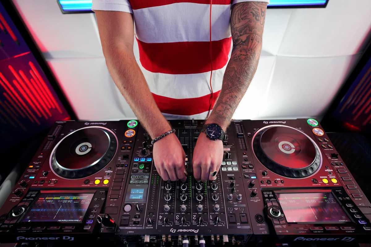 A guy standing behind a DJ desk with many buttons and knobs.